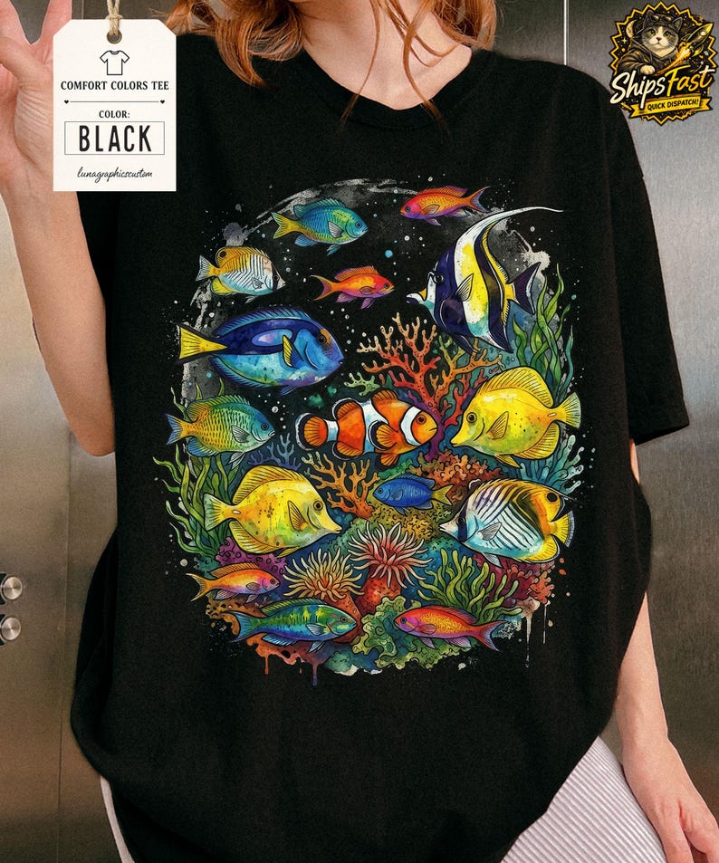 May include: Black Comfort Colors tee featuring a vibrant, colorful illustration of various tropical fish swimming around coral and seaweed. The shirt includes a tag that says "BLACK".