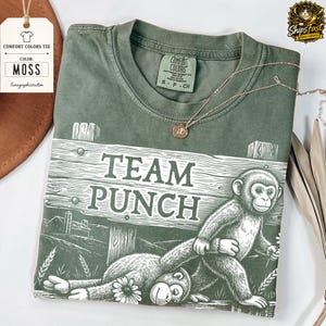 May include: A moss green Comfort Colors tee shirt featuring the text "TEAM PUNCH" and a graphic of two monkeys. A gold necklace is draped across the shirt. The shirt has a tag that says "MOSS".