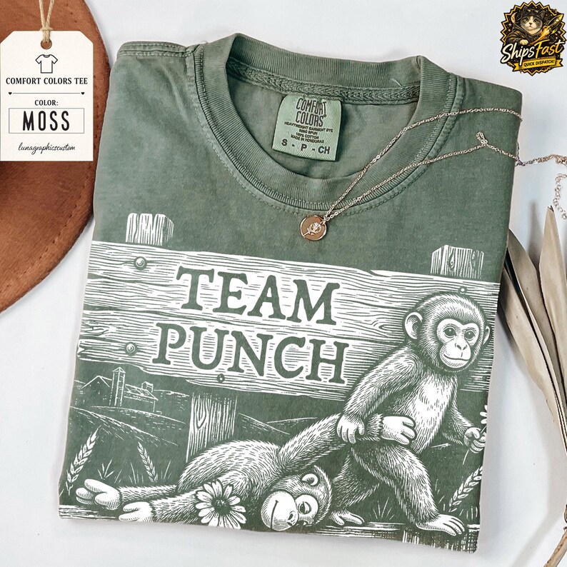 May include: A moss green Comfort Colors tee shirt featuring the text "TEAM PUNCH" and a graphic of two monkeys. A gold necklace is draped across the shirt. The shirt has a tag that says "MOSS".