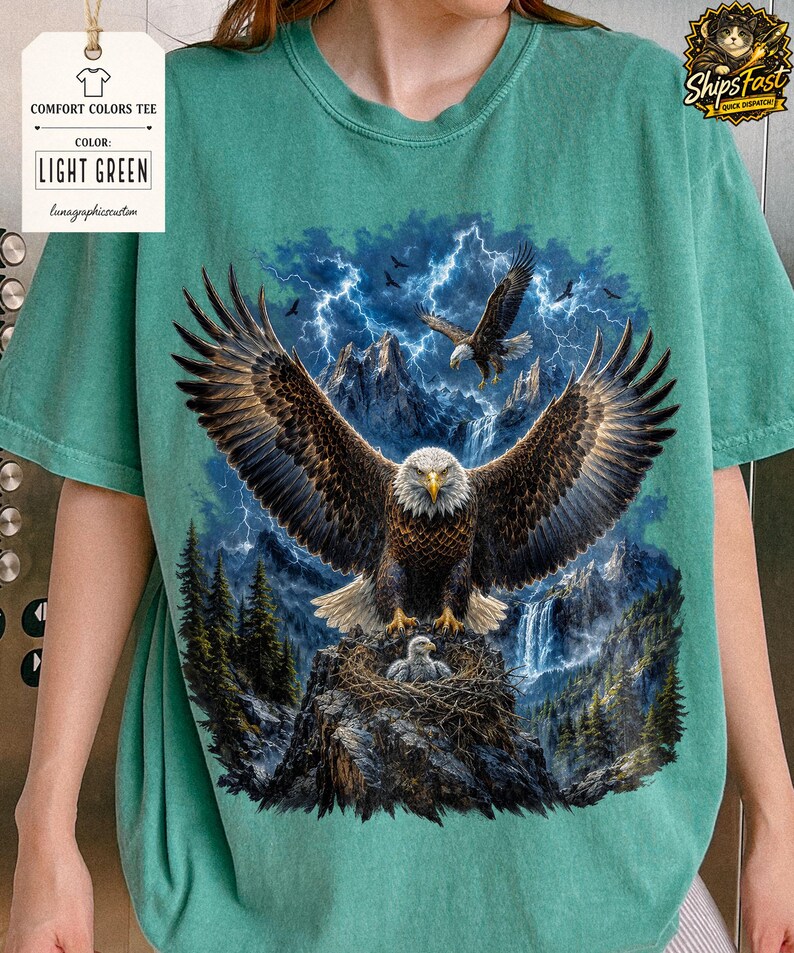 May include: Light green Comfort Colors tee featuring a detailed graphic of eagles with outstretched wings, a nest with an eaglet, and a mountain backdrop with lightning. The shirt includes the text "LIGHT GREEN".