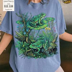 May include: Blue jean Comfort Colors tee featuring a detailed illustration of two green frogs in a pond setting. The frogs are on lily pads and logs, surrounded by aquatic plants and flowers. The shirt has the text "BLUE JEAN".