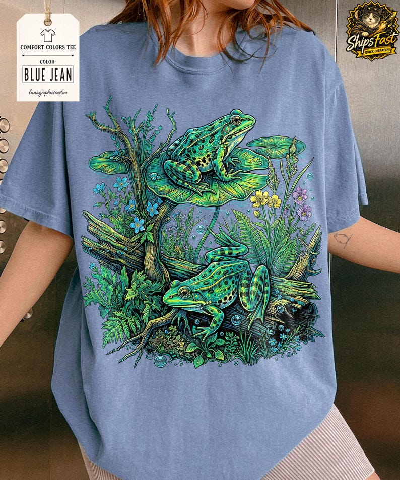 May include: Blue jean Comfort Colors tee featuring a detailed illustration of two green frogs in a pond setting. The frogs are on lily pads and logs, surrounded by aquatic plants and flowers. The shirt has the text "BLUE JEAN".