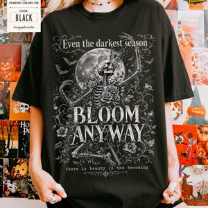 May include: A black t-shirt featuring a white graphic print of a skeleton surrounded by roses, vines, and a full moon. The text reads: Even the darkest season, BLOOM ANYWAY, there is beauty in the becoming. The design includes small bats and a butterfly.