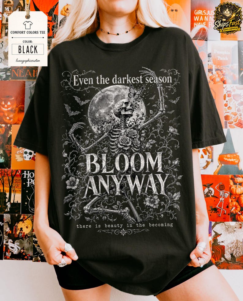 May include: A black t-shirt featuring a white graphic print of a skeleton surrounded by roses, vines, and a full moon. The text reads: Even the darkest season, BLOOM ANYWAY, there is beauty in the becoming. The design includes small bats and a butterfly.