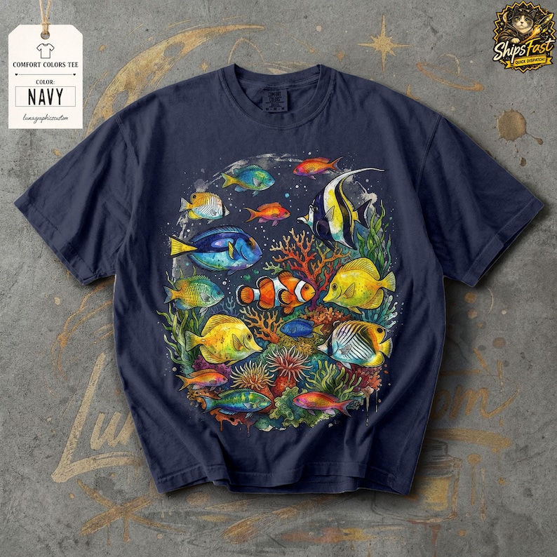 May include: Navy blue Comfort Colors tee featuring a colorful illustration of various fish species swimming around coral. The design includes a clownfish, angelfish, and other vibrant marine life. The tag reads "COMFORT COLORS TEE" and "NAVY".