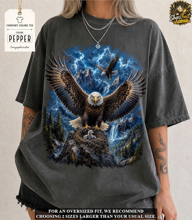 May include: Pepper-colored Comfort Colors tee featuring a detailed graphic of a bald eagle with outstretched wings, perched on a nest with eaglets. The design includes a mountain backdrop, lightning, and text. The shirt is oversized.