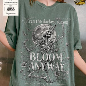 May include: A moss green t-shirt featuring a graphic print of a skeleton surrounded by roses, a butterfly, and a full moon. The text reads: Even the darkest season, BLOOM ANYWAY, there is beauty in the becoming. The design is in shades of white and gray.