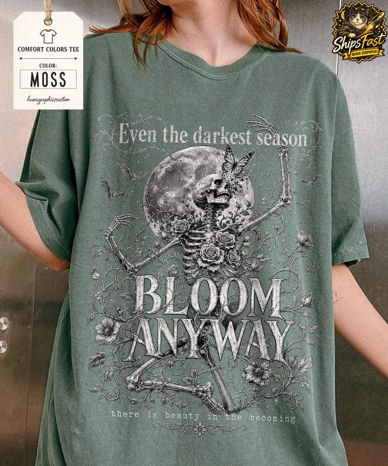May include: A moss green t-shirt featuring a graphic print of a skeleton surrounded by roses, a butterfly, and a full moon. The text reads: Even the darkest season, BLOOM ANYWAY, there is beauty in the becoming. The design is in shades of white and gray.