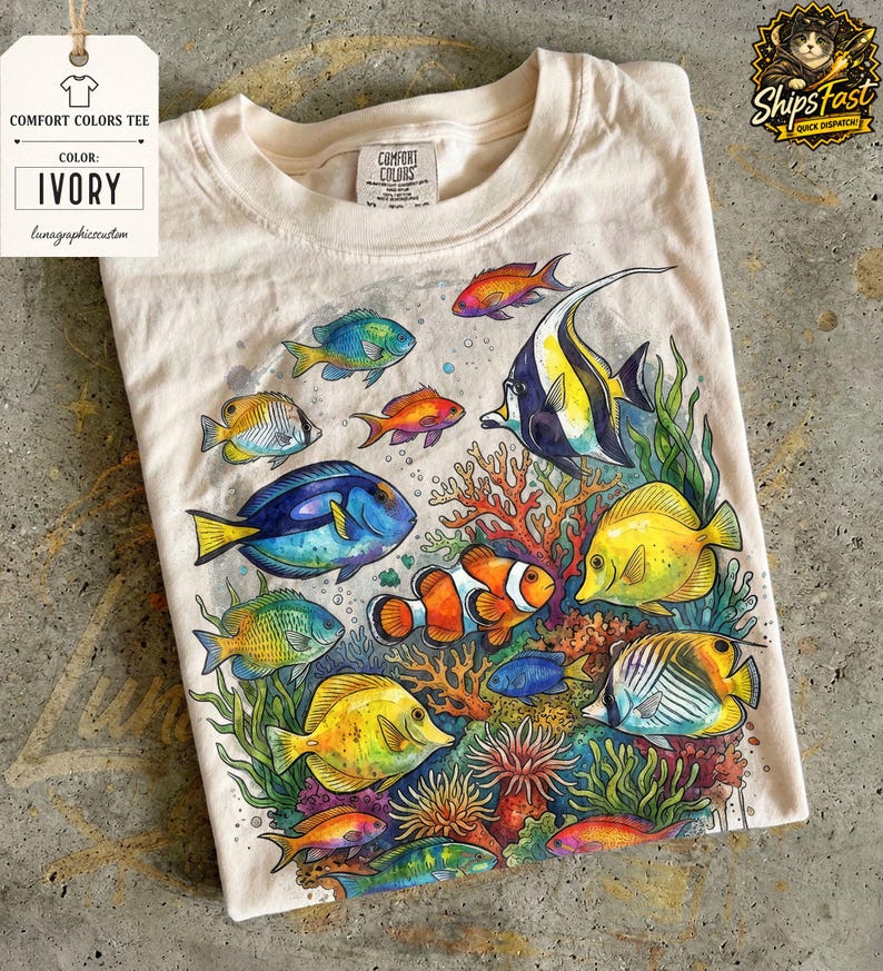 May include: Ivory Comfort Colors tee featuring a vibrant watercolor illustration of various colorful fish and coral. The design includes a clownfish, blue tang, and other tropical fish. The shirt has a tag that says "Comfort Colors Tee" and "Ivory".