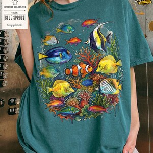 May include: Blue spruce Comfort Colors tee featuring a colorful illustration of various fish swimming around coral. The design includes a clownfish, a blue tang, and other tropical fish. The shirt has a relaxed fit.