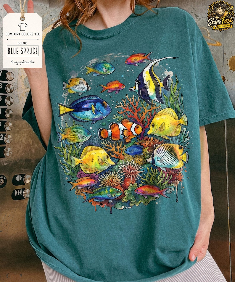 May include: Blue spruce Comfort Colors tee featuring a colorful illustration of various fish swimming around coral. The design includes a clownfish, a blue tang, and other tropical fish. The shirt has a relaxed fit.