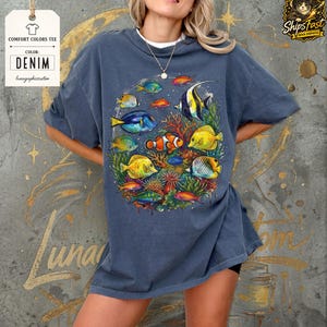 May include: Denim blue Comfort Colors tee featuring a vibrant underwater scene with colorful fish and coral. The graphic includes various fish species in shades of yellow, blue, orange, and red. The tee has the text "Comfort Colors Tee" and "Denim".