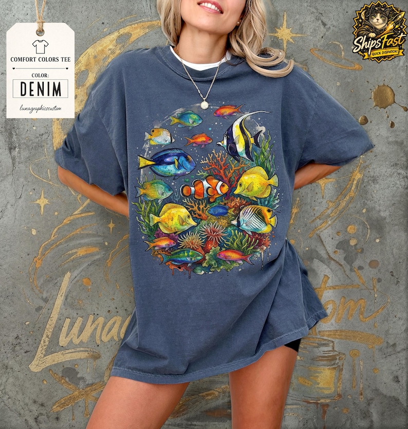 May include: Denim blue Comfort Colors tee featuring a vibrant underwater scene with colorful fish and coral. The graphic includes various fish species in shades of yellow, blue, orange, and red. The tee has the text "Comfort Colors Tee" and "Denim".