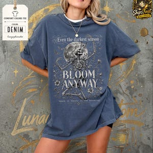 May include: A denim blue short-sleeve t-shirt featuring a graphic print of a skeleton surrounded by flowers, a moon, and butterflies. The text reads: Even the darkest season BLOOM ANYWAY there is beauty in the becoming. The shirt has a crew neckline.