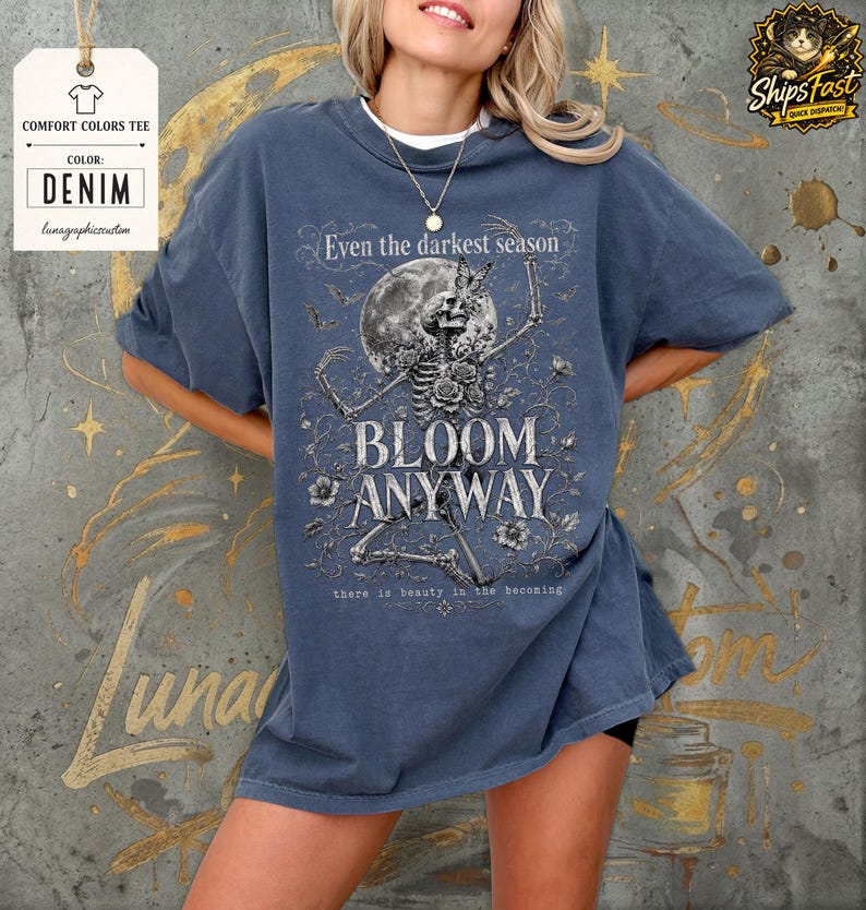 May include: A denim blue short-sleeve t-shirt featuring a graphic print of a skeleton surrounded by flowers, a moon, and butterflies. The text reads: Even the darkest season BLOOM ANYWAY there is beauty in the becoming. The shirt has a crew neckline.