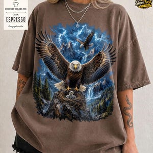 May include: Espresso-colored Comfort Colors tee featuring a detailed eagle graphic with outstretched wings, set against a mountain and lightning backdrop. The design includes an eagle in flight and a nest with eaglets. The shirt has a vintage, worn look.