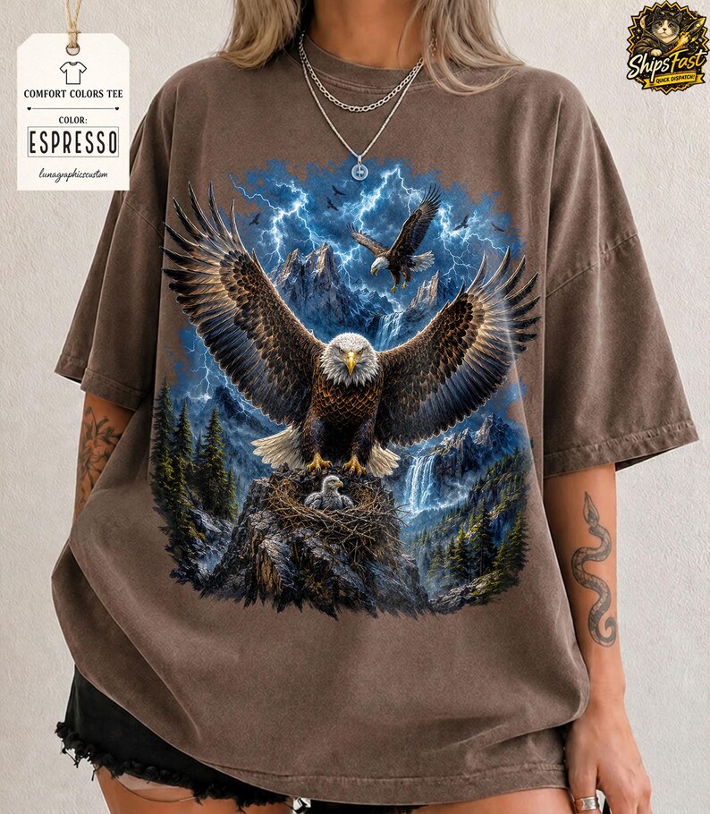 May include: Espresso-colored Comfort Colors tee featuring a detailed eagle graphic with outstretched wings, set against a mountain and lightning backdrop. The design includes an eagle in flight and a nest with eaglets. The shirt has a vintage, worn look.