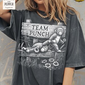 May include: Dark grey t-shirt featuring a black and white illustration of two monkeys on a wooden fence. The sign on the fence reads "TEAM PUNCH". The design includes flowers and grass.