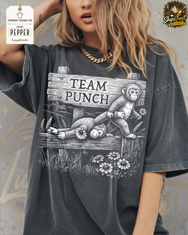 May include: Dark grey t-shirt featuring a black and white illustration of two monkeys on a wooden fence. The sign on the fence reads "TEAM PUNCH". The design includes flowers and grass.