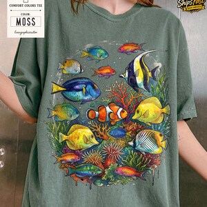 May include: Moss green Comfort Colors tee featuring a vibrant underwater scene. The graphic showcases colorful fish, coral, and aquatic plants. The shirt has the text "Comfort Colors Tee" and "Moss" on a tag.