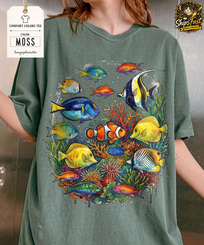 May include: Moss green Comfort Colors tee featuring a vibrant underwater scene. The graphic showcases colorful fish, coral, and aquatic plants. The shirt has the text "Comfort Colors Tee" and "Moss" on a tag.