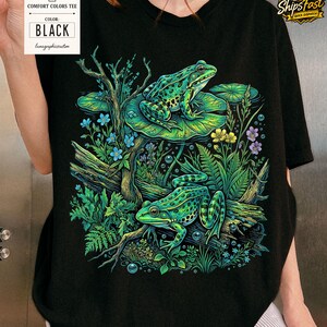May include: Black Comfort Colors tee featuring a vibrant illustration of two green frogs amidst lush foliage, including lily pads, flowers, and branches. The shirt has a tag that says "Comfort Colors Tee" and "Black".