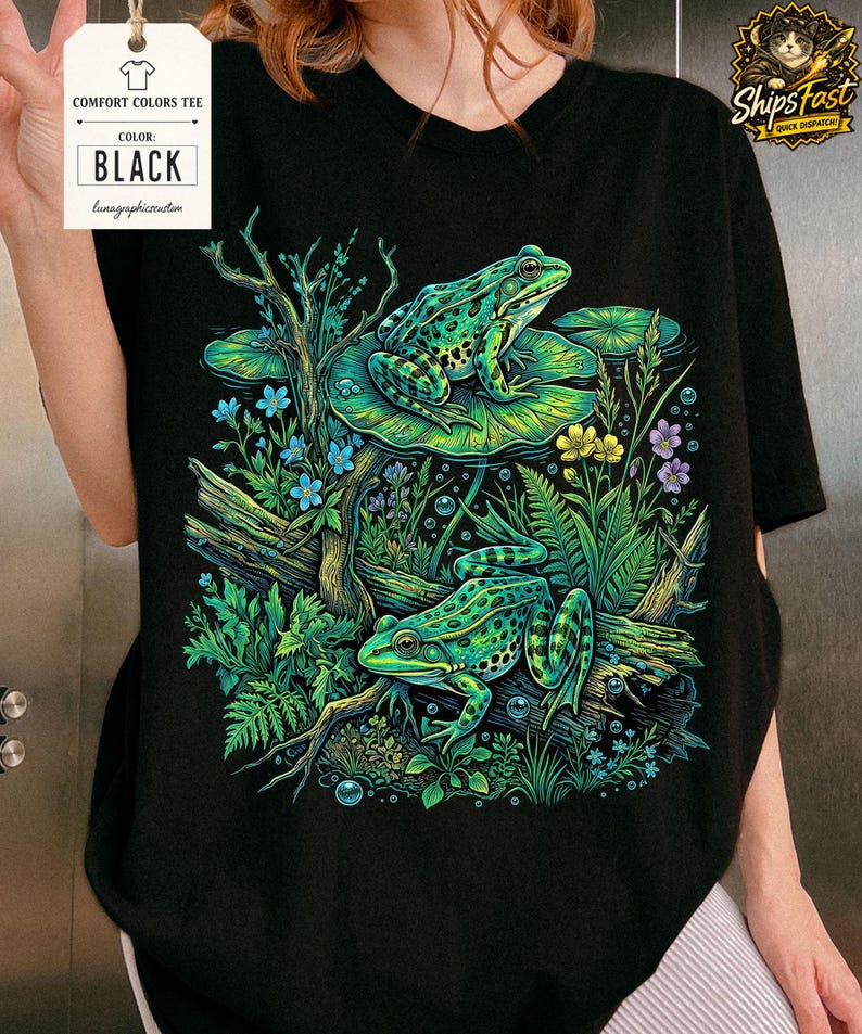 May include: Black Comfort Colors tee featuring a vibrant illustration of two green frogs amidst lush foliage, including lily pads, flowers, and branches. The shirt has a tag that says "Comfort Colors Tee" and "Black".