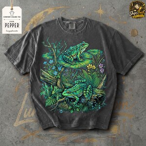 May include: Dark gray Comfort Colors tee featuring a vibrant illustration of two green frogs amidst lush aquatic plants and flowers. The design includes lily pads, logs, and bubbles, creating a detailed nature scene. The tag reads "Comfort Colors Tee" and "Pepper".