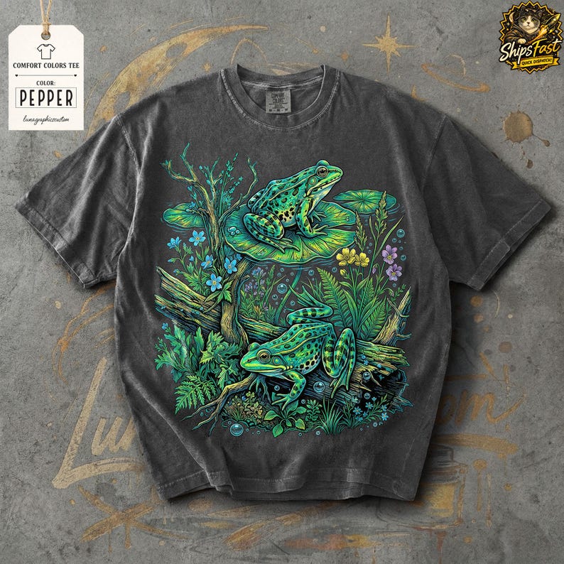 May include: Dark gray Comfort Colors tee featuring a vibrant illustration of two green frogs amidst lush aquatic plants and flowers. The design includes lily pads, logs, and bubbles, creating a detailed nature scene. The tag reads "Comfort Colors Tee" and "Pepper".