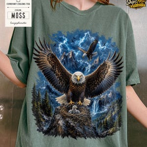 May include: Moss green Comfort Colors tee featuring a detailed graphic of eagles soaring over a mountain landscape with a waterfall and lightning. The design includes an eagle in a nest with a baby eagle. The text "MOSS" is displayed.