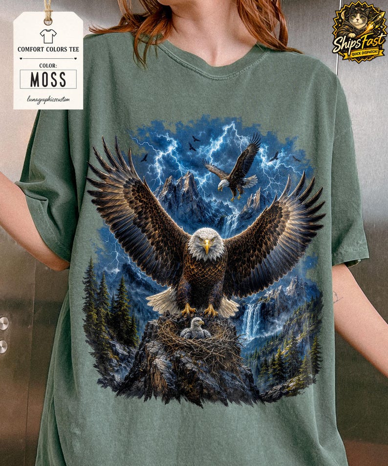 May include: Moss green Comfort Colors tee featuring a detailed graphic of eagles soaring over a mountain landscape with a waterfall and lightning. The design includes an eagle in a nest with a baby eagle. The text "MOSS" is displayed.