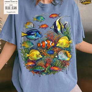 May include: Blue jean Comfort Colors tee featuring a vibrant underwater scene. The design showcases colorful fish, coral, and aquatic plants. The shirt includes the text "BLUE JEAN" on a tag.