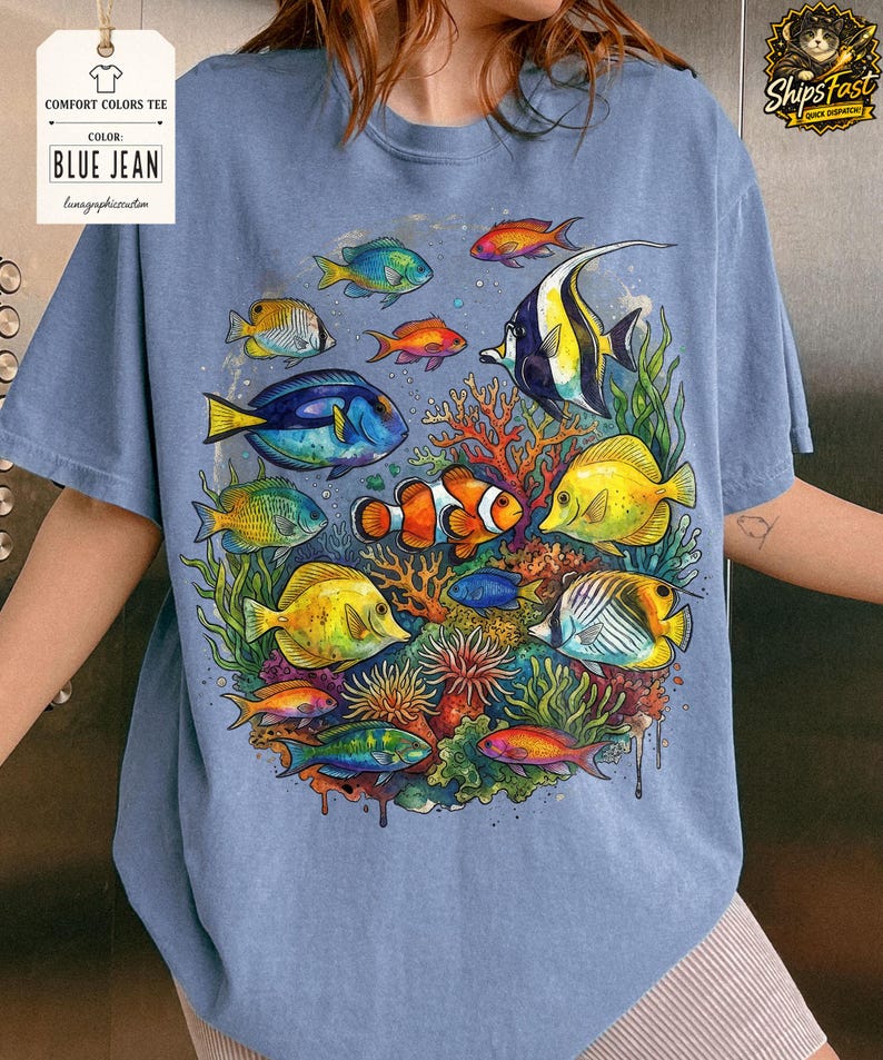 May include: Blue jean Comfort Colors tee featuring a vibrant underwater scene. The design showcases colorful fish, coral, and aquatic plants. The shirt includes the text "BLUE JEAN" on a tag.