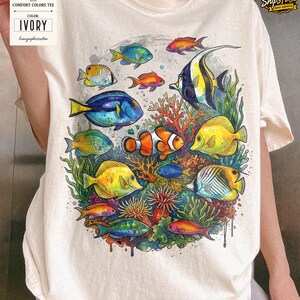 May include: Ivory Comfort Colors tee featuring a vibrant watercolor design of various colorful fish and coral. The design includes fish in shades of blue, yellow, orange, and green, creating an underwater scene. The tee has a relaxed fit.