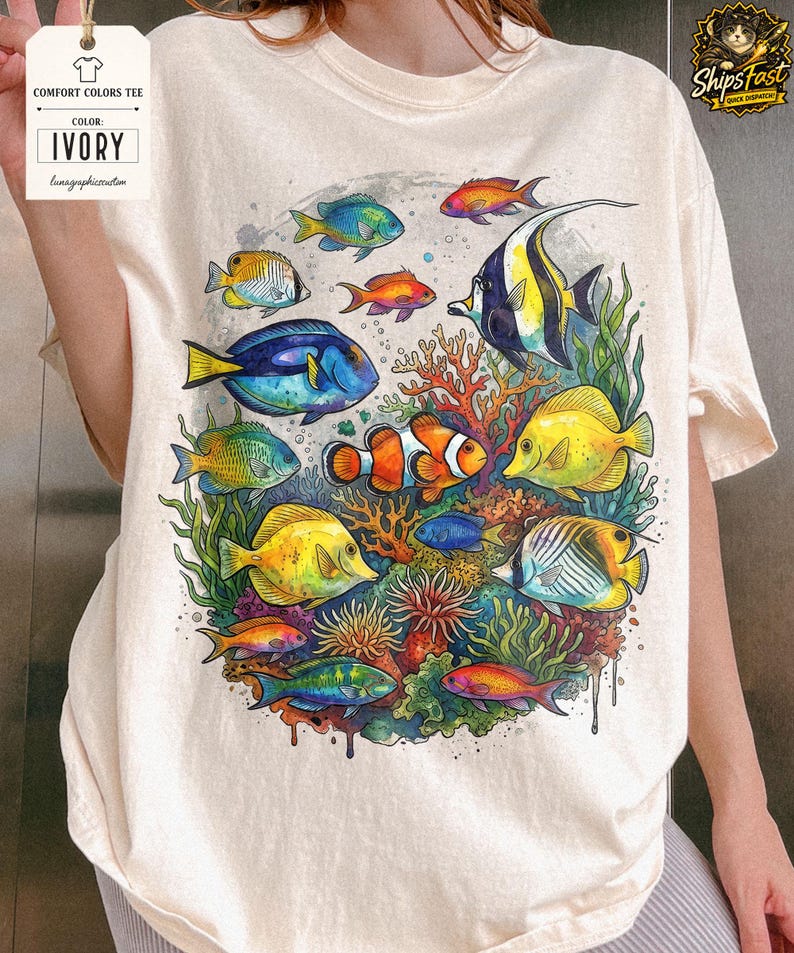 May include: Ivory Comfort Colors tee featuring a vibrant watercolor design of various colorful fish and coral. The design includes fish in shades of blue, yellow, orange, and green, creating an underwater scene. The tee has a relaxed fit.