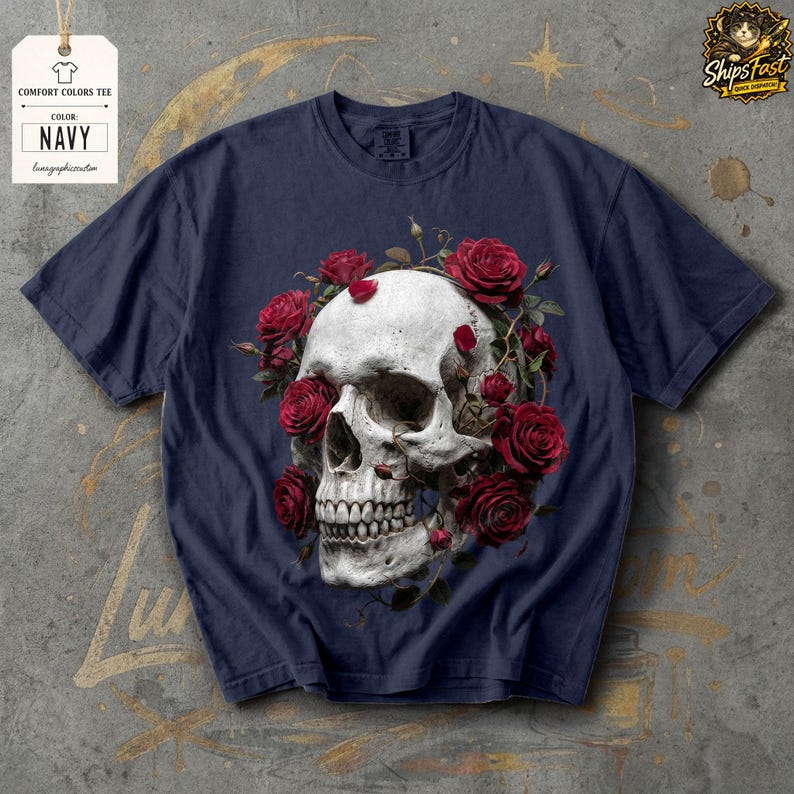 May include: Navy blue comfort colors tee featuring a detailed skull design encircled by vibrant red roses. The shirt includes a tag with the text "COMFORT COLORS TEE" and "NAVY". The design is centered on the front of the shirt.
