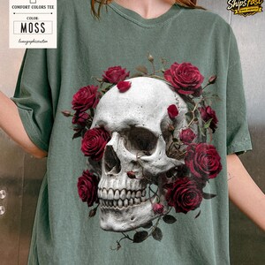 May include: A moss green Comfort Colors tee featuring a detailed skull design encircled by vibrant red roses. The skull has a realistic appearance, and the roses add a contrasting pop of color. The shirt has the text "Comfort Colors Tee" and "Color: Moss".