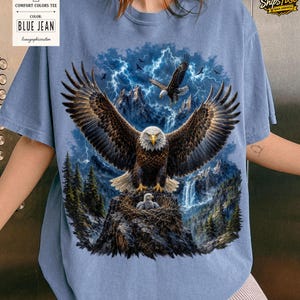 May include: Blue jean Comfort Colors tee featuring a detailed graphic of an eagle with outstretched wings, a nest with eaglets, and a mountain backdrop with lightning. The shirt has the text "BLUE JEAN" on a tag.