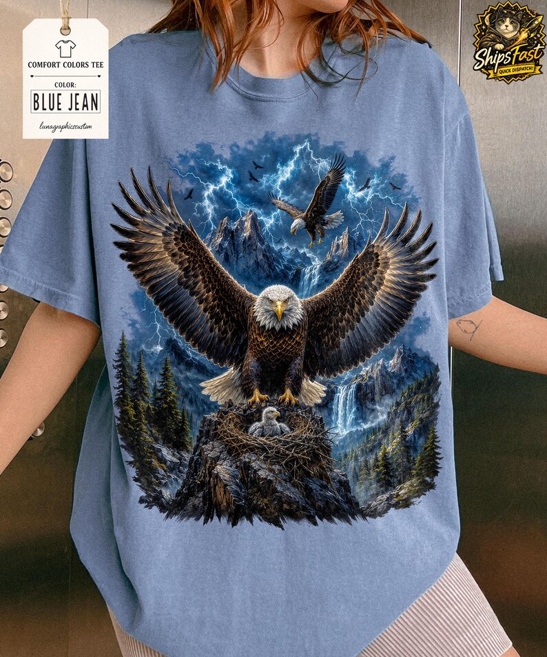 May include: Blue jean Comfort Colors tee featuring a detailed graphic of an eagle with outstretched wings, a nest with eaglets, and a mountain backdrop with lightning. The shirt has the text "BLUE JEAN" on a tag.