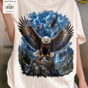 May include: Ivory Comfort Colors tee featuring a detailed graphic of a bald eagle with outstretched wings, perched on a nest with an eaglet. The background includes mountains, a waterfall, and a stormy sky with lightning and other eagles.