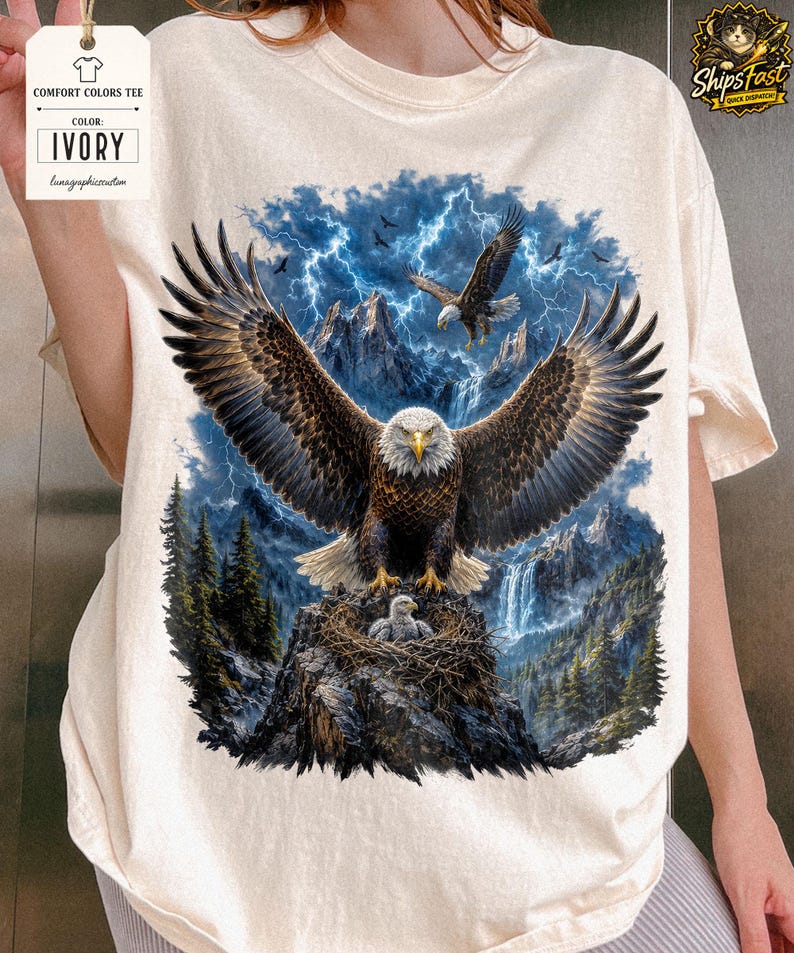May include: Ivory Comfort Colors tee featuring a detailed graphic of a bald eagle with outstretched wings, perched on a nest with an eaglet. The background includes mountains, a waterfall, and a stormy sky with lightning and other eagles.