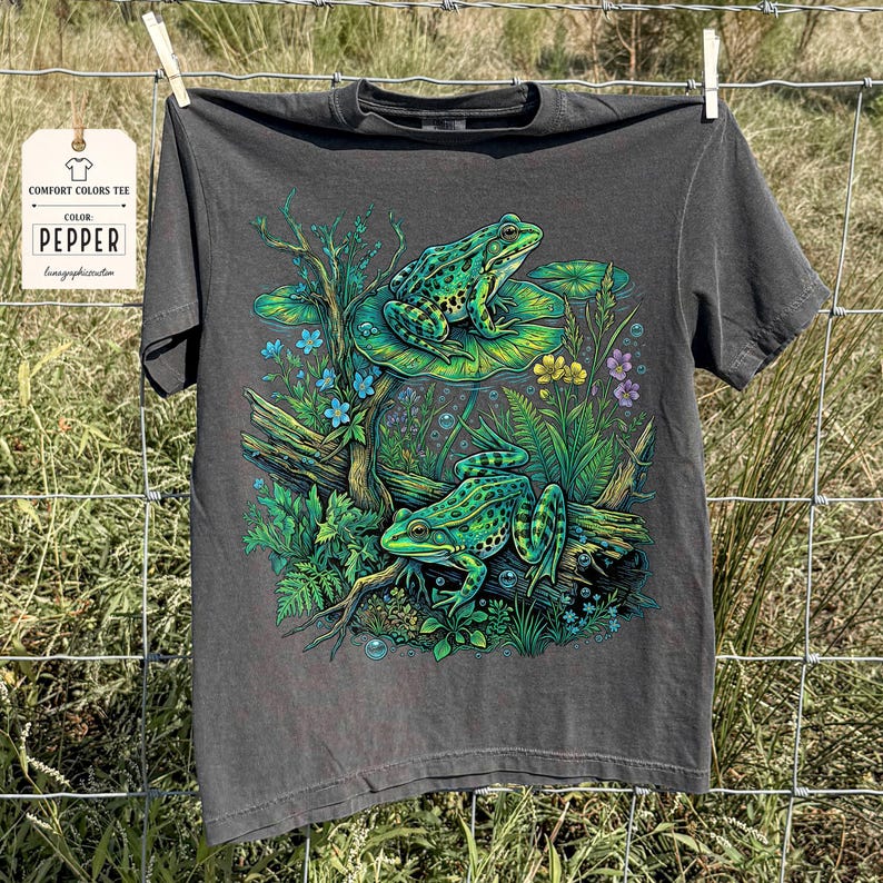 May include: A dark gray Comfort Colors T-shirt with a vibrant illustration of two green frogs in a pond setting. The frogs are on lily pads and a log, surrounded by plants and flowers. The tag reads "Pepper".