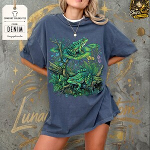 May include: Denim blue Comfort Colors tee featuring a vibrant illustration of two green frogs on a lily pad and log, surrounded by lush greenery and flowers. The tee has the text "Lunar Graphic Creation" on a tag.