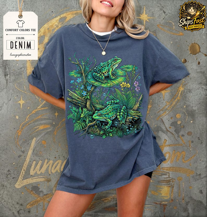 May include: Denim blue Comfort Colors tee featuring a vibrant illustration of two green frogs on a lily pad and log, surrounded by lush greenery and flowers. The tee has the text "Lunar Graphic Creation" on a tag.