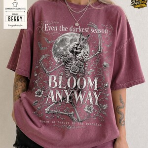 May include: A berry-colored short-sleeve t-shirt featuring a graphic print of a skeleton surrounded by roses, a butterfly, and a full moon. The text reads: Even the darkest season, BLOOM ANYWAY, there is beauty in the becoming.