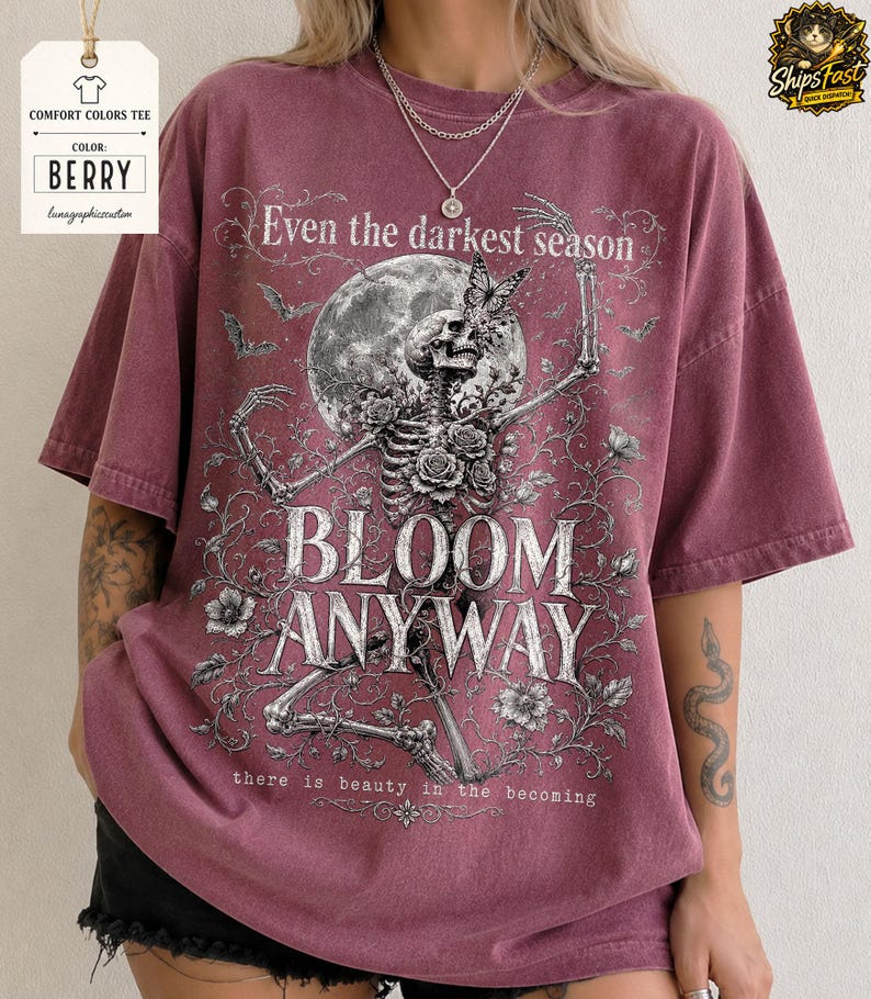 May include: A berry-colored short-sleeve t-shirt featuring a graphic print of a skeleton surrounded by roses, a butterfly, and a full moon. The text reads: Even the darkest season, BLOOM ANYWAY, there is beauty in the becoming.