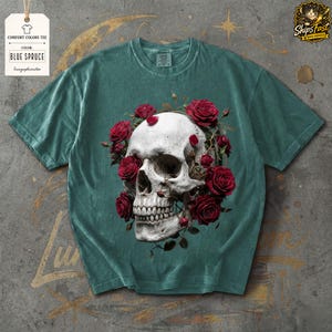 May include: Blue spruce comfort colors tee featuring a detailed skull design encircled by red roses. The graphic tee includes a tag with the text "Comfort Colors Tee" and "Blue Spruce".