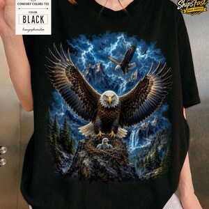 May include: Black Comfort Colors tee featuring a detailed graphic of eagles soaring over a mountain landscape with a waterfall and lightning. The shirt includes a tag that says "Comfort Colors Tee" and "Black".