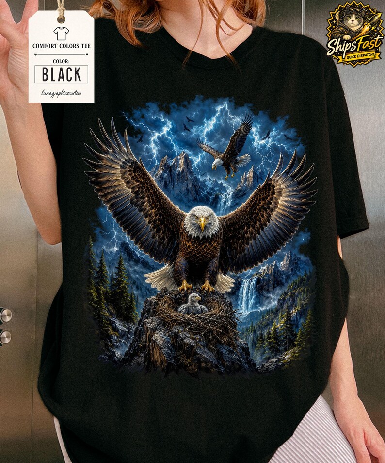 May include: Black Comfort Colors tee featuring a detailed graphic of eagles soaring over a mountain landscape with a waterfall and lightning. The shirt includes a tag that says "Comfort Colors Tee" and "Black".