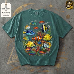 May include: Blue spruce comfort colors tee featuring a colorful illustration of various tropical fish, including clownfish, angelfish, and butterflyfish, swimming around coral. The shirt has a crew neck and short sleeves. The tag reads "Comfort Colors Tee" and "Blue Spruce".